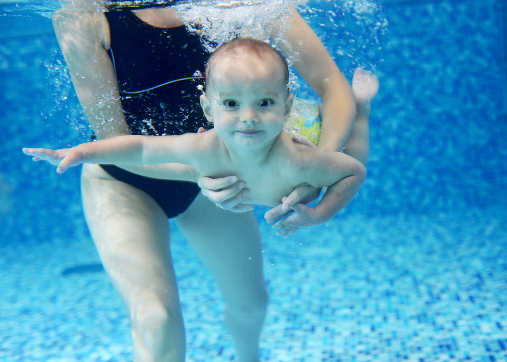 Toddler boy learning to swim under water with the support of his mom
